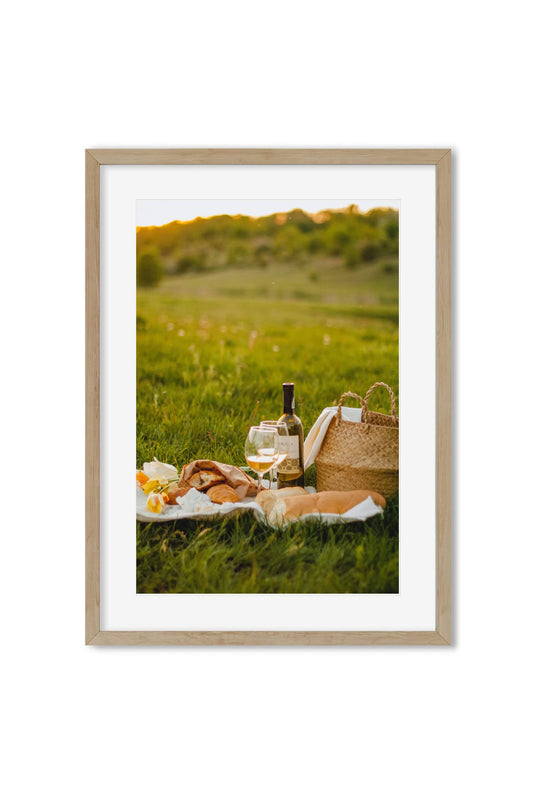 Golden-hour picnic scene with wine, bread, flowers and a wicker basket in a green meadow