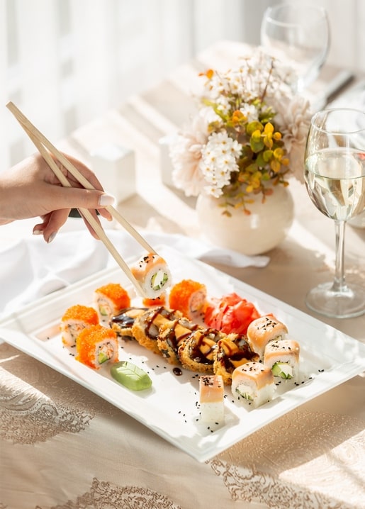 Hand hebt mit Stäbchen eine Sushi-Rolle von einer Platte neben Blumen und Wein