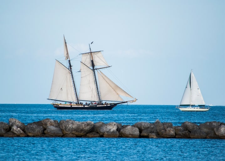 Segelschiff mit vollen Segeln fährt an einer Steinmole vorbei, kleines Segelboot dahinter
