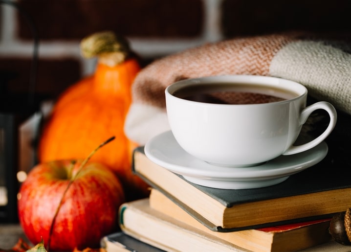 Cup of tea on stacked books with a pumpkin, red apple and plaid blanket