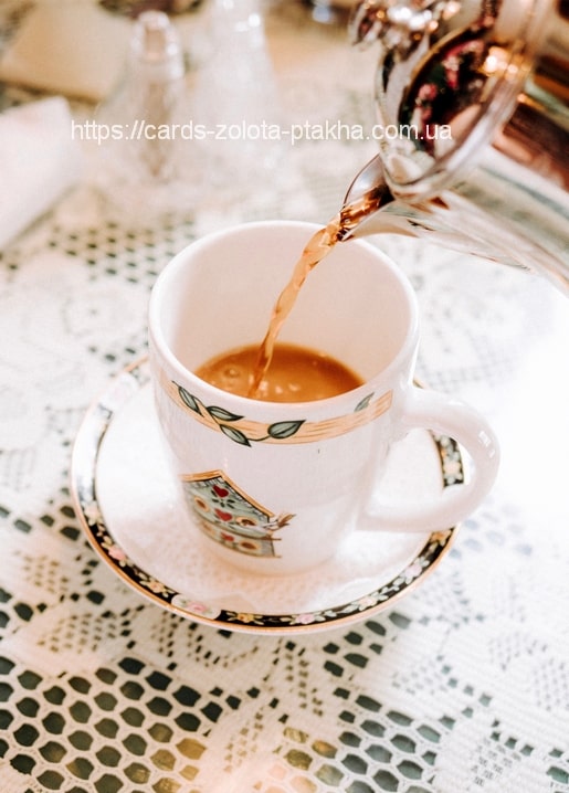 A silver teapot pours tea into a floral-patterned teacup on a lace tablecloth.