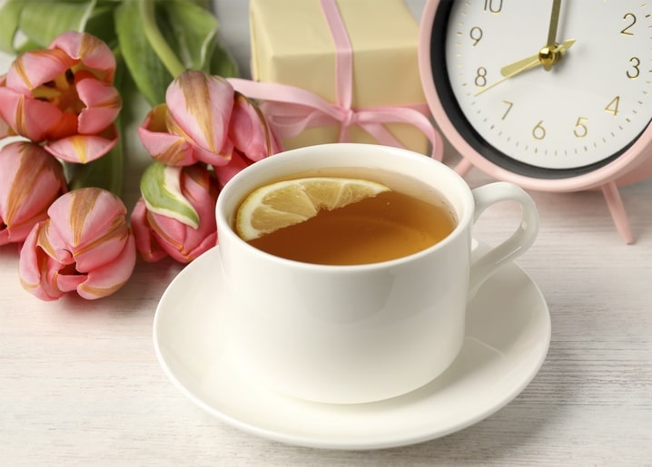 White teacup with lemon tea, pink tulips, a ribboned gift box and a pastel alarm clock