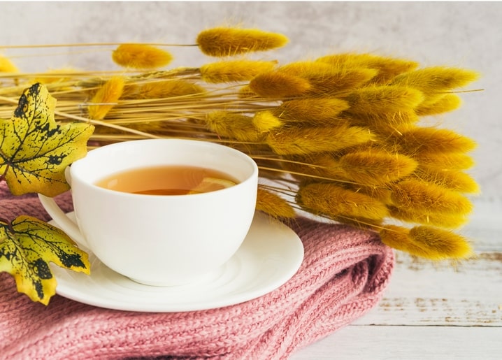 White teacup on pink knit fabric with golden bunny-tail grass and spotted leaves