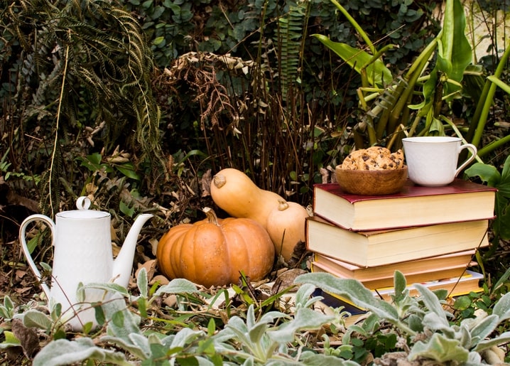 White teapot, a pumpkin and butternut squashes, and a stack of books with coffee and cookies in a garden