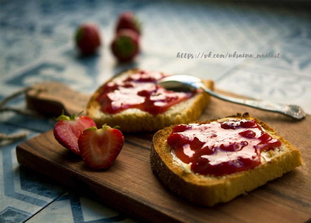 Toast spread with strawberry jam beside fresh strawberries on a wooden board