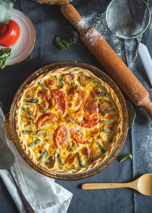Golden quiche topped with tomato and green beans beside a rolling pin and flour sieve on dark linen.