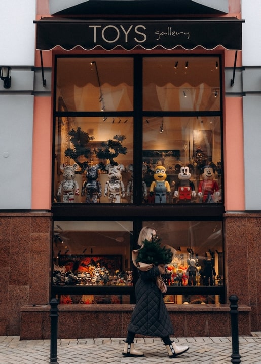 Woman carrying pine branches walks past a snowy toy shop window filled with collectible figures