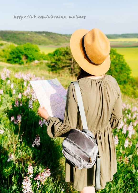 Frau mit Strohhut hält eine Papierkarte in einer Blumenwiese vor grünen Hügeln