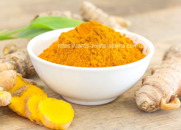 Bowl of golden ground turmeric beside fresh turmeric roots and a green leaf on a wood table