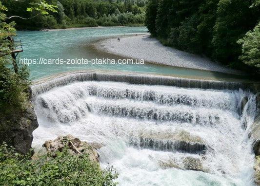 Türkisfarbener Fluss stürzt über ein breites Wehr nahe einem bewaldeten Kiesstrand