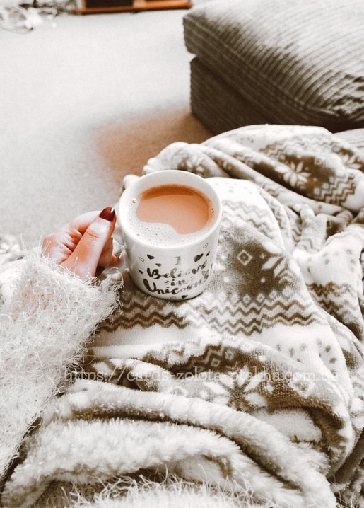 Hand im flauschigen Pullover hält eine Einhorn-Tasse mit Kakao auf einer Decke mit Schneeflockenmuster