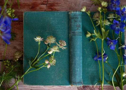 Verwittertes petrolfarbenes Buch auf einem Holztisch, umgeben von blauen und weißen Wildblumen