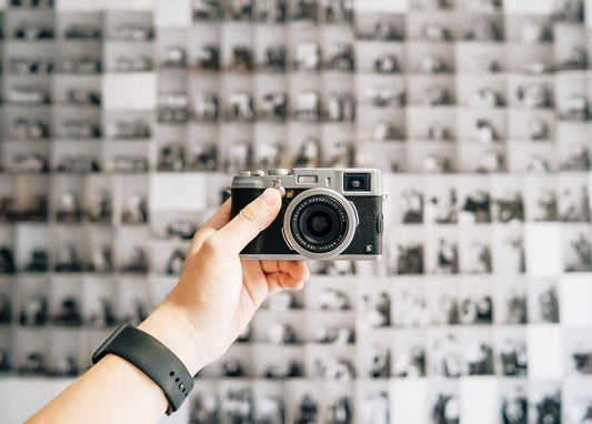 Hand hält eine Retro-Kamera vor einer Wand aus Schwarz-Weiß-Kontaktabzügen
