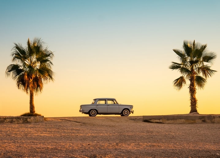 Vintage light-blue sedan on a desert road flanked by two tall palm trees at sunset