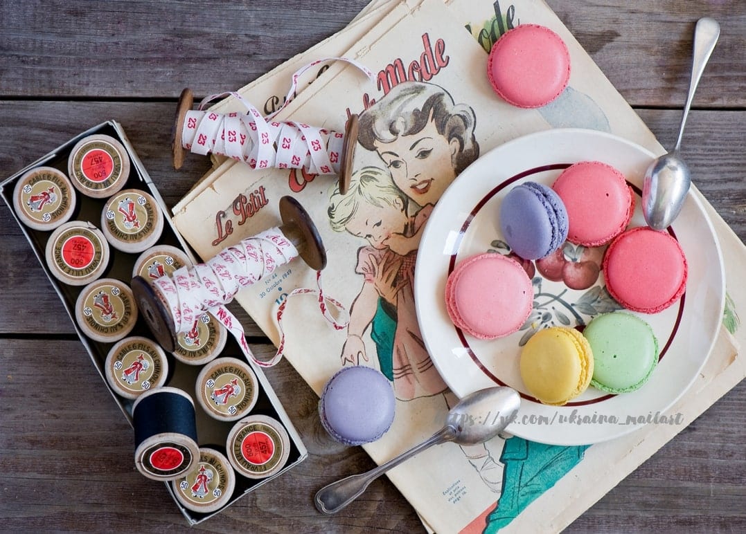 Hölzerne Garnrollen, Bandspulen, eine alte französische Modezeitschrift und ein Teller pastellfarbener Macarons
