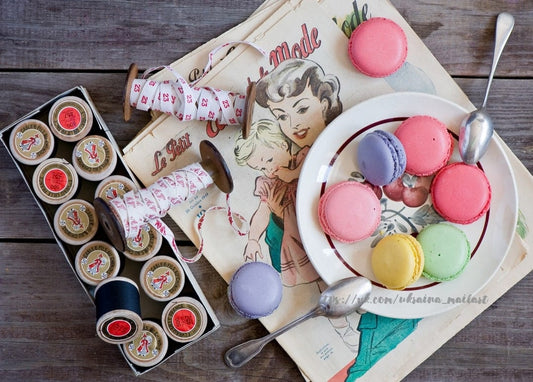 Hölzerne Garnrollen, Bandspulen, eine alte französische Modezeitschrift und ein Teller pastellfarbener Macarons