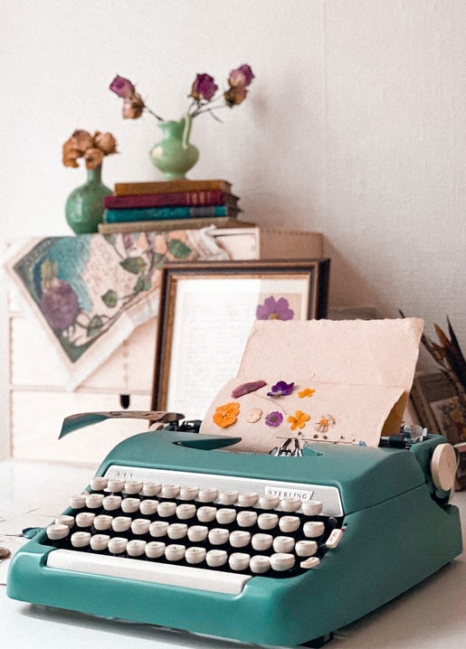 Türkisfarbene Vintage-Schreibmaschine mit gepressten Stiefmütterchen, Trockenrosen und Büchern