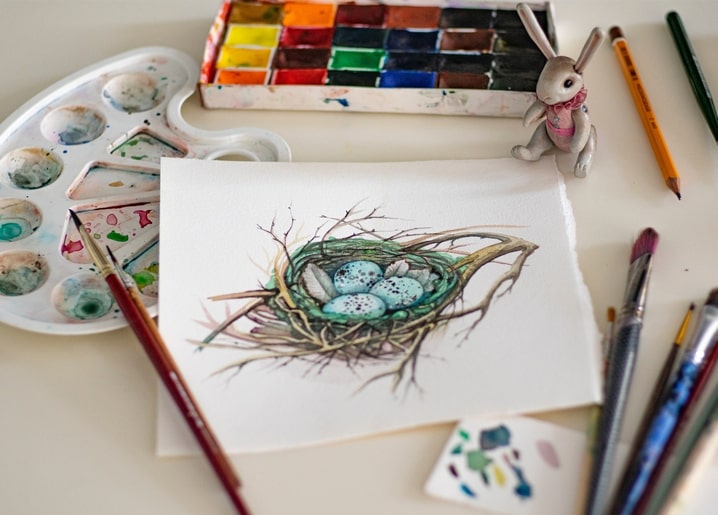 Aquarellmalerei eines Vogelnests mit gesprenkelten Eiern neben Farbpalette und Pinseln