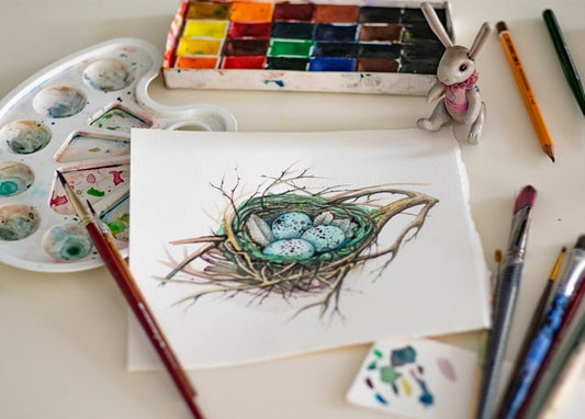 Watercolor painting of a bird's nest with speckled blue eggs beside a paint palette and brushes