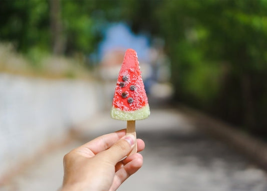 Hand hält ein wassermelonenförmiges Eis am Stiel vor sonniger Straße
