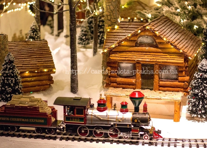 Miniatur-Zug White Christmas Express fährt an schneebedeckten Blockhütten mit Lichterketten vorbei