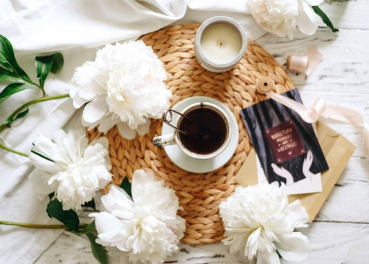 Weiße Pfingstrosen um eine Tasse Kaffee, eine brennende Kerze und eine Geschenkkarte mit Schleife