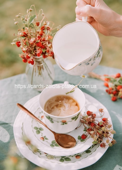 Tee wird in eine mit Walderdbeeren bemalte Tasse gegossen, daneben Beeren und ein goldener Löffel
