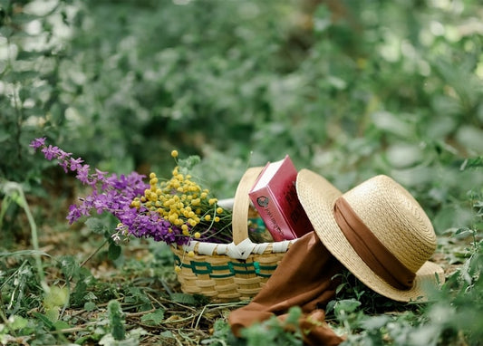 Geflochtener Korb mit Wiesenblumen, Buch und Strohhut im Gras
