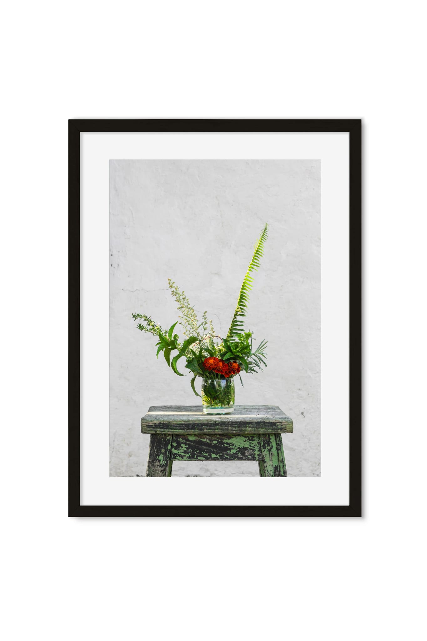 Red wildflowers and ferns in a glass jar on a weathered green stool against a white wall