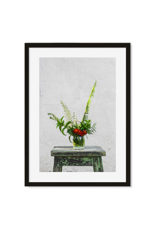 Red wildflowers and ferns in a glass jar on a weathered green stool against a white wall