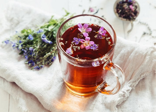 Glass mug of herbal tea with pink wildflowers, beside a lavender bouquet and tea strainer on linen