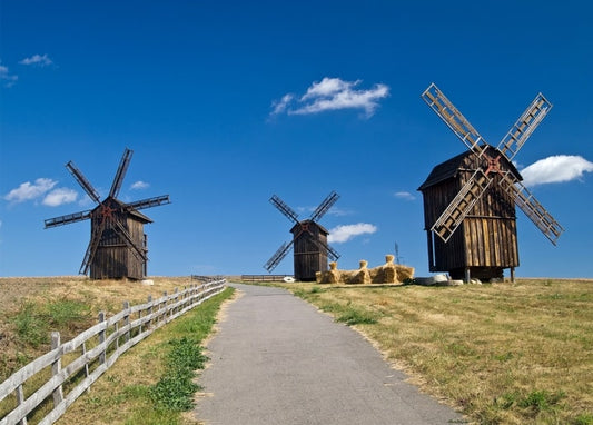 Три дерев'яні вітряки з розкритими крилами вздовж польової стежки під синім небом, поряд — тюки сіна.