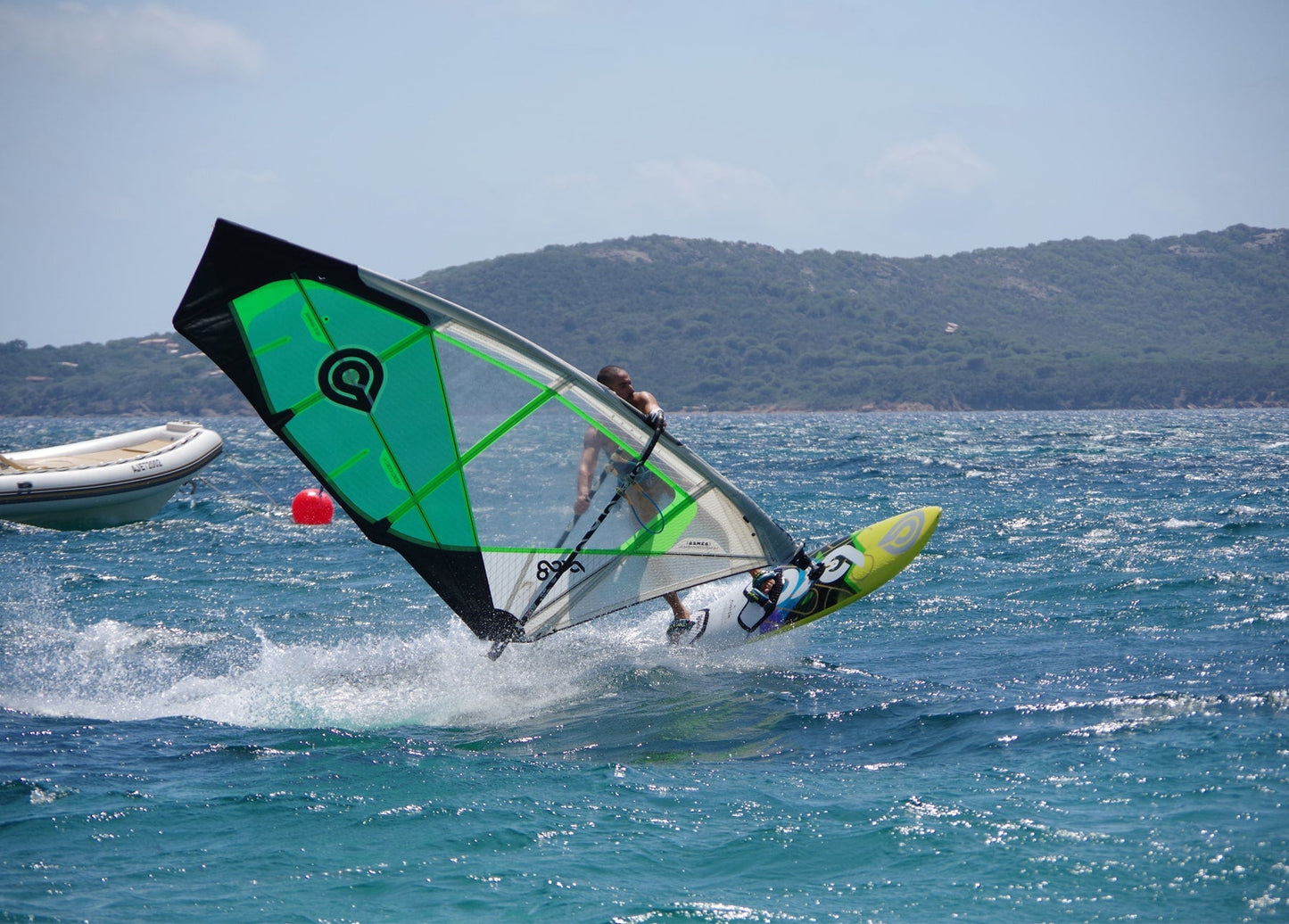 Windsurfer mit grün-schwarzem Segel springt über eine Welle vor einer hügeligen Küste