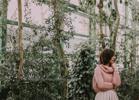 Frau im rosa Strickpullover zwischen grünen Pflanzen in einem Glasgewächshaus