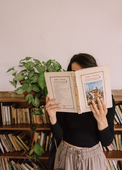 Дівчина ховає обличчя за книгою Жуля Верна біля книжкової полиці з вазоном