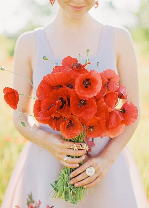 Frau im hellen Kleid hält einen großen Strauß roter Mohnblumen auf einem sonnigen Feld