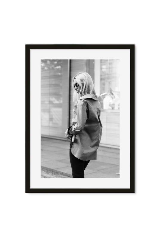 Black-and-white street-style photo of a woman in an oversized trench coat glancing back on a city street