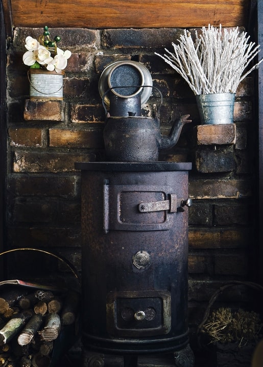 Antique black wood stove with a kettle on top, brick wall, dried flowers, and firewood basket