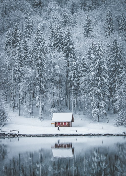 Small lit wooden cabin on a snowy lakeshore, reflected in still water