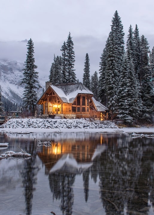 Timber cabin glowing at dusk beside a still winter lake surrounded by snowy pines