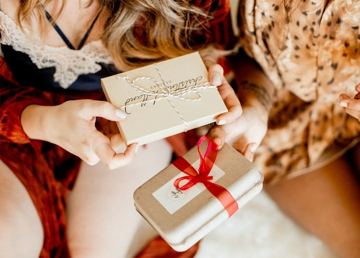 Zwei Personen tauschen kleine Geschenkboxen, gebunden mit Kordel und roter Schleife.