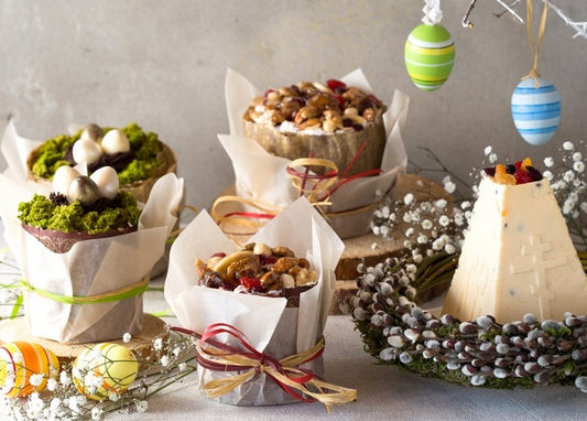 Small paska cakes wrapped in craft paper with moss and eggs, framed by a pussy willow wreath and hanging painted eggs.