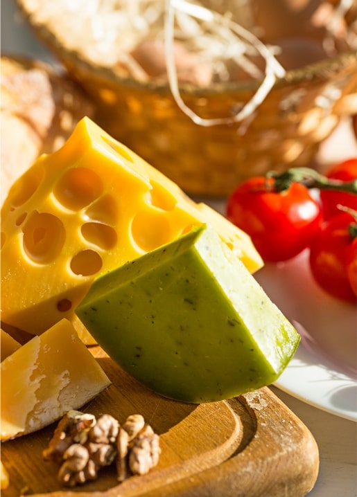 Holey yellow cheese and green herb cheese wedges beside cherry tomatoes and a bread basket