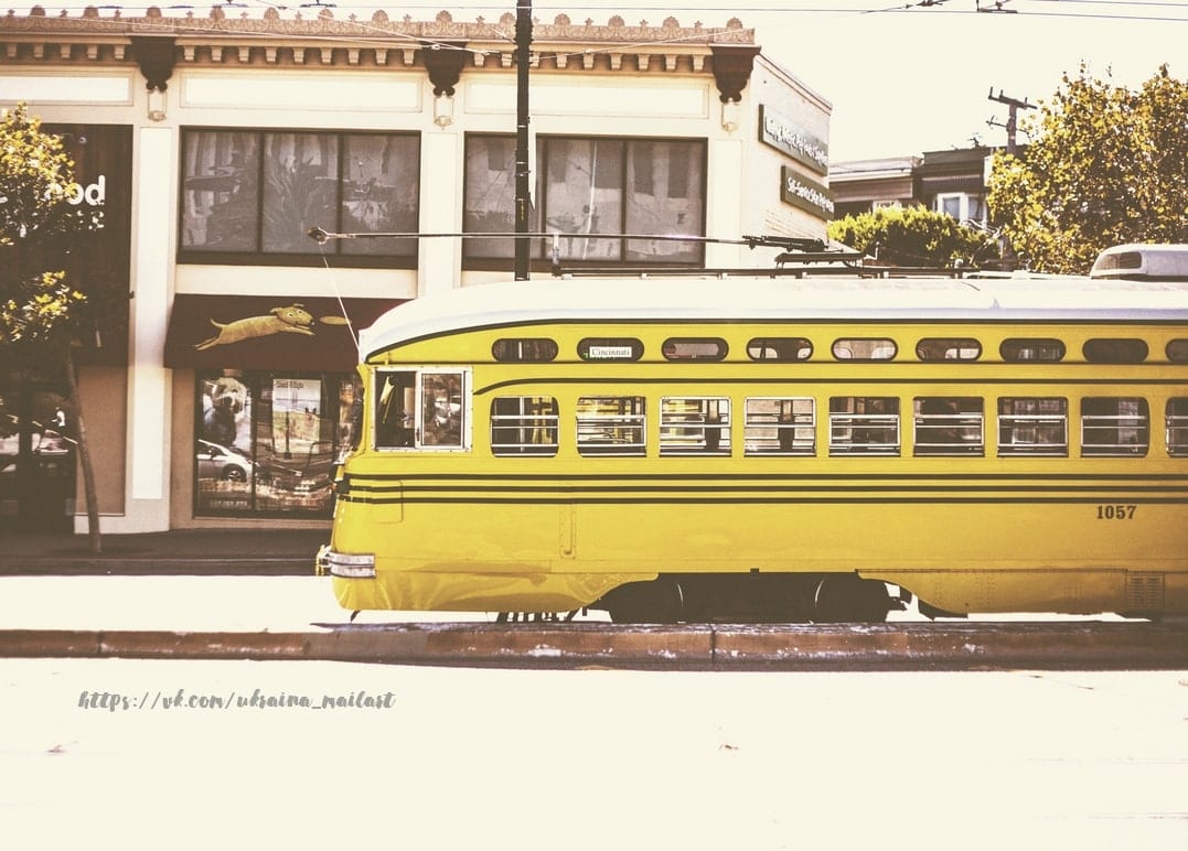 Gelbe Vintage-Straßenbahn fährt an Schaufenstern auf einer Stadtstraße vorbei